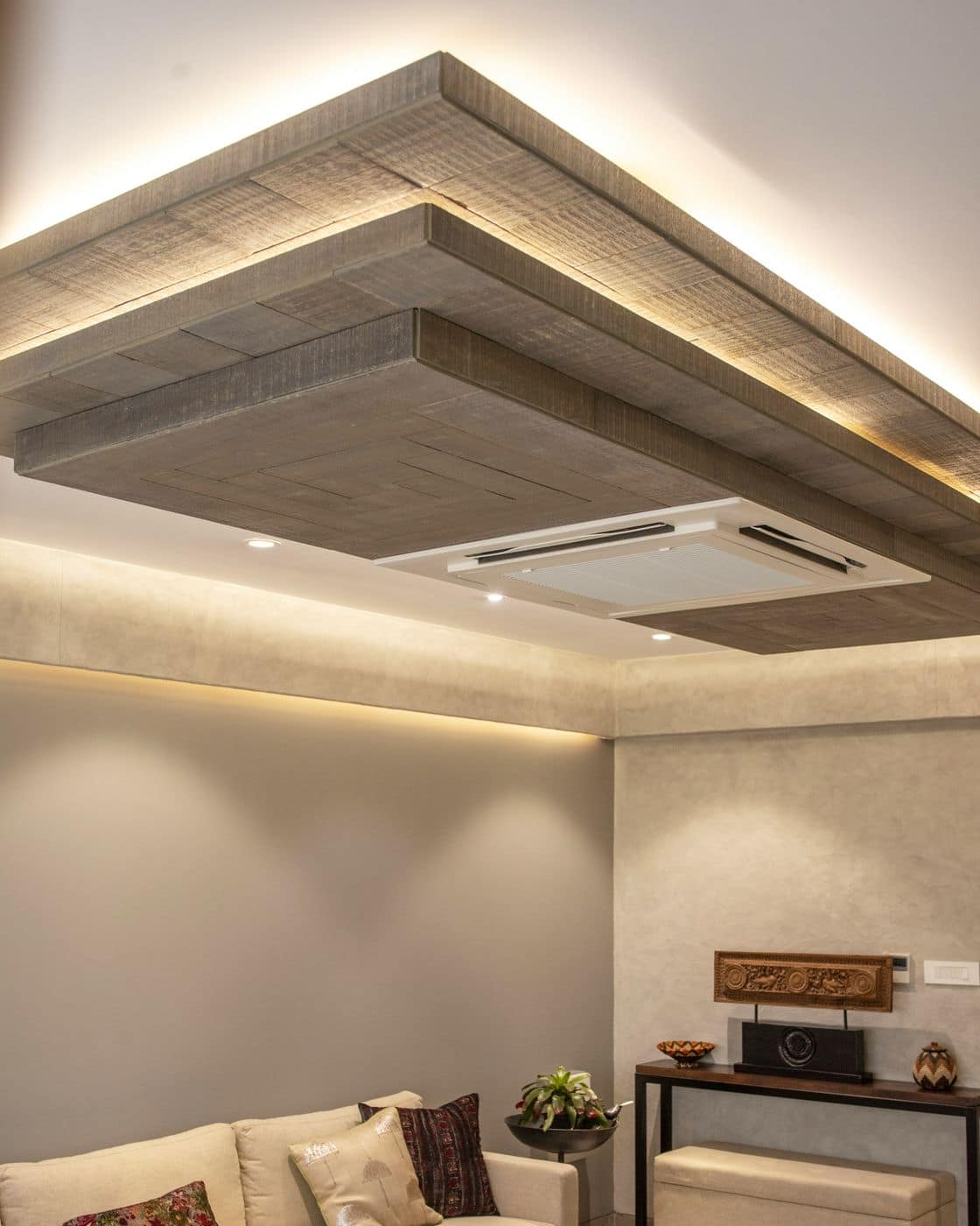 Layered false ceiling with wood-finish tray, warm cove lighting and cassette AC above a living area