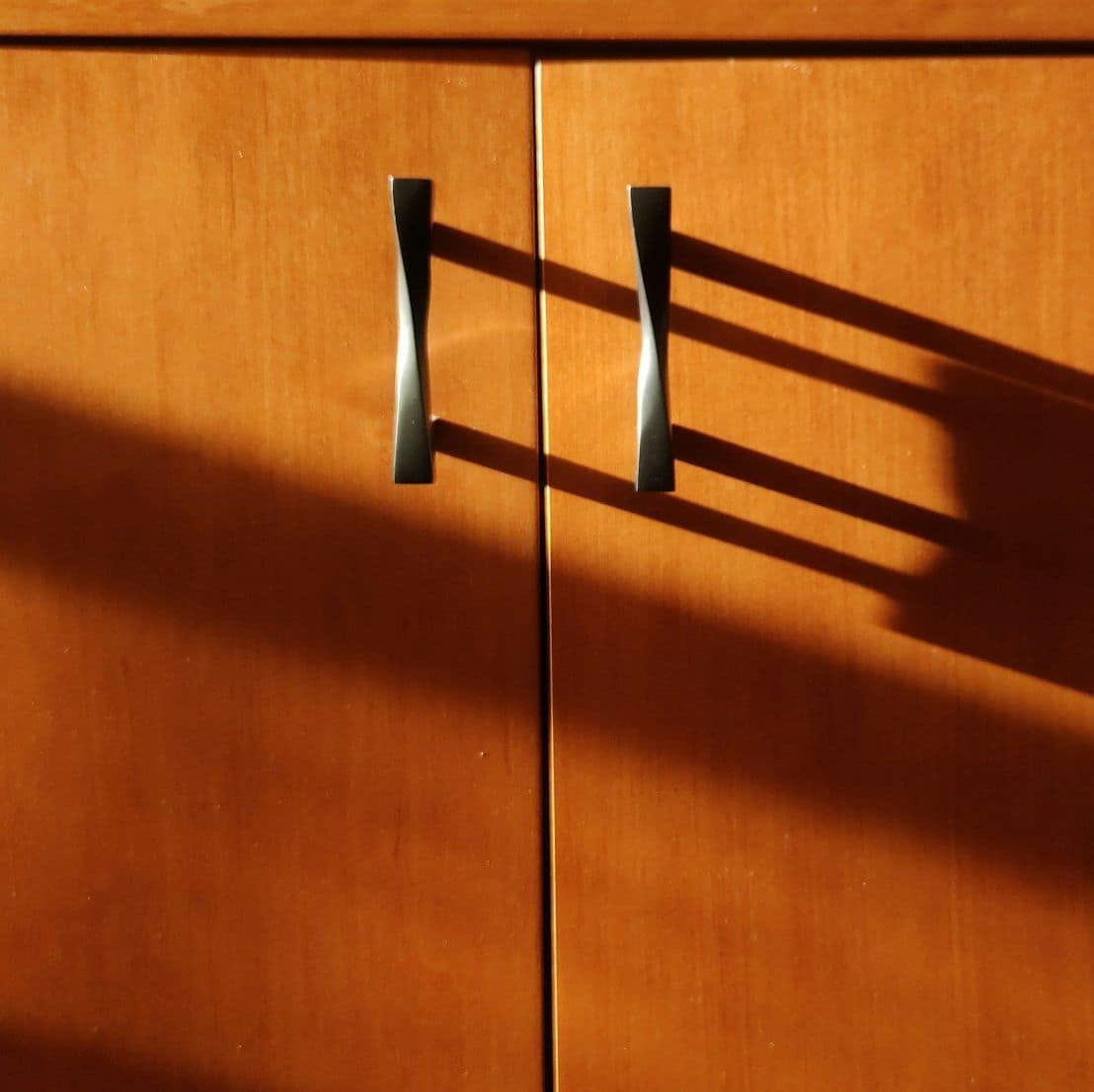 Two twisted metal pull handles on honey-toned wooden cabinet doors with strong sunlit shadows