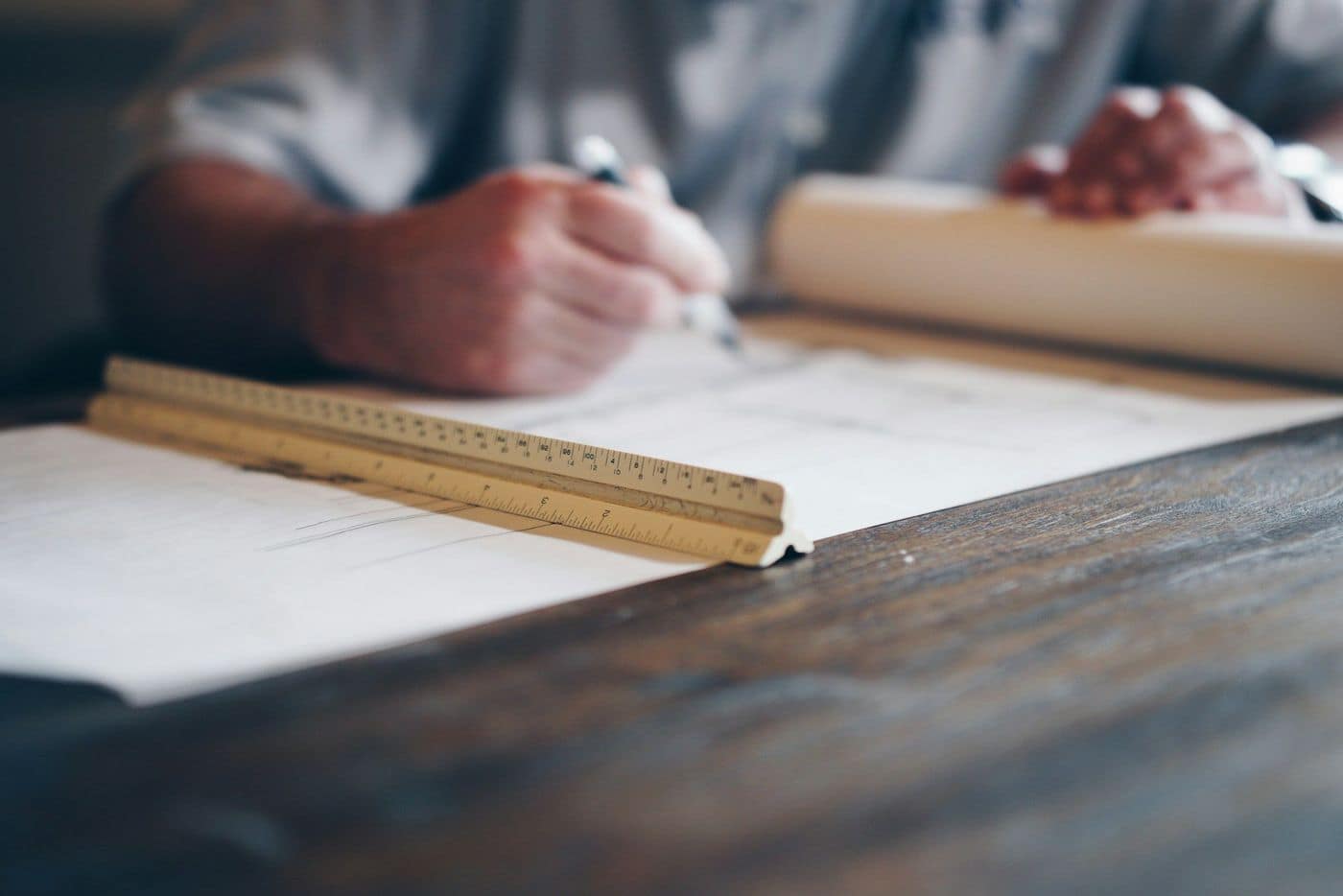 Hands drafting on a paper plan with a triangular scale ruler on a dark wooden table
