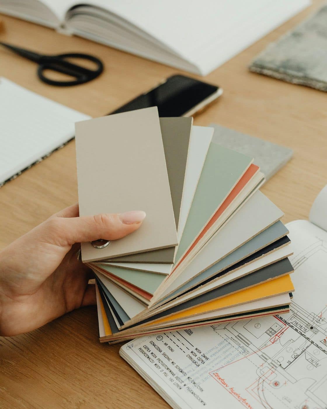 Hand holding a fan of neutral colour swatches over floor plans and an open book on a wooden desk