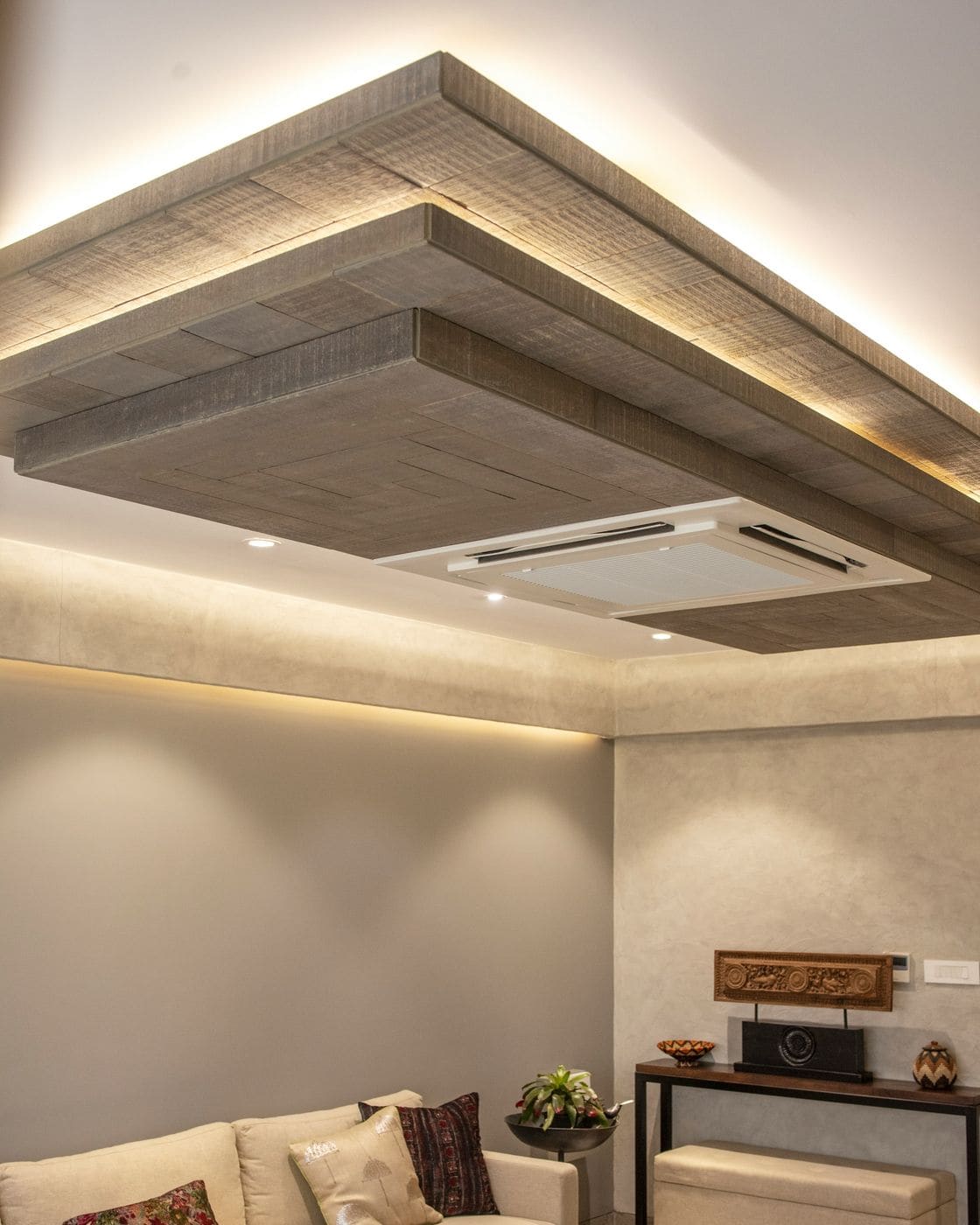 Layered false ceiling with wood-finish tray, warm cove lighting and cassette AC above a living area