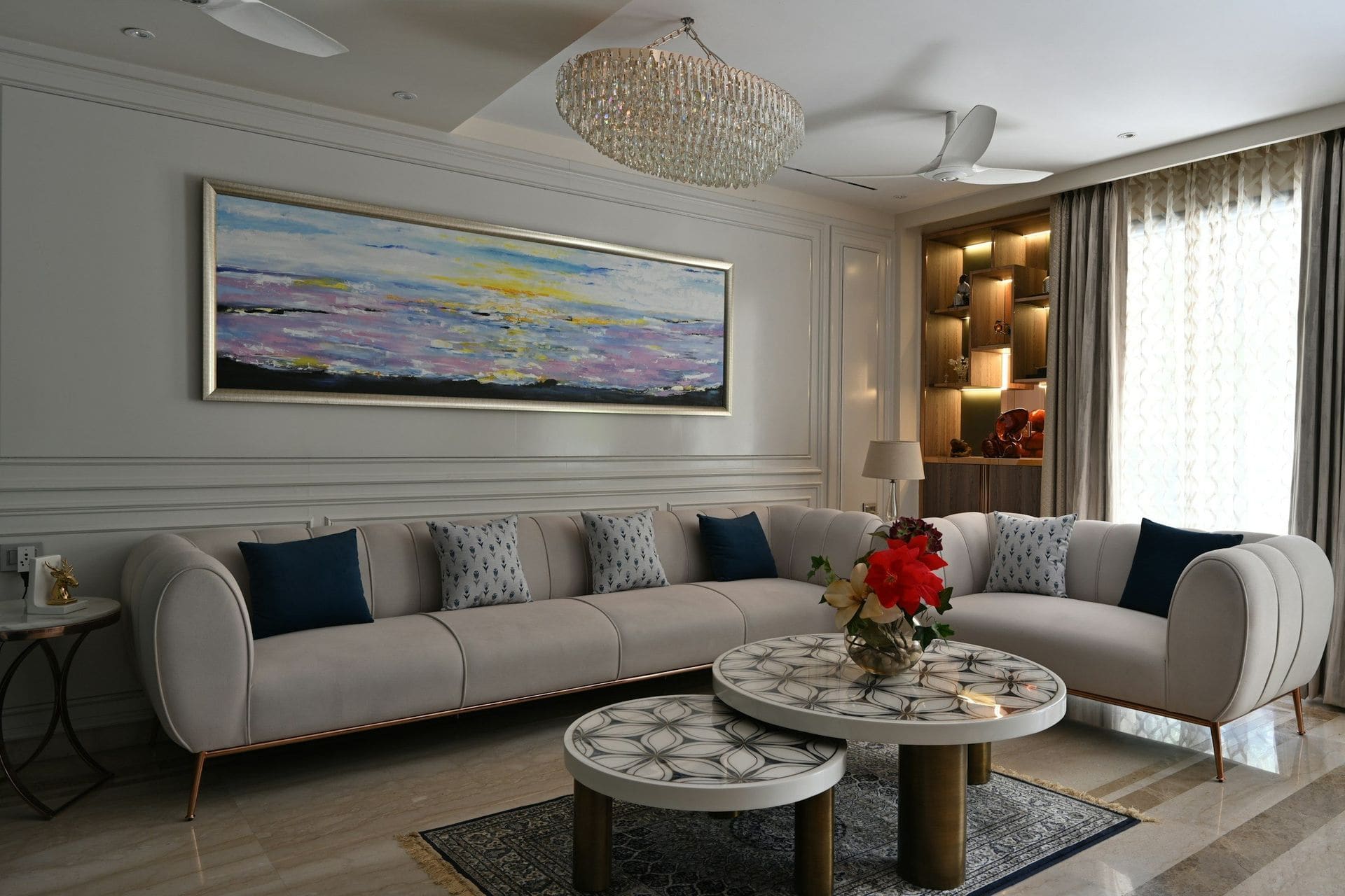 Indian living room with grey sectional sofa, navy cushions, panelled ivory walls, crystal chandelier and ceiling fans