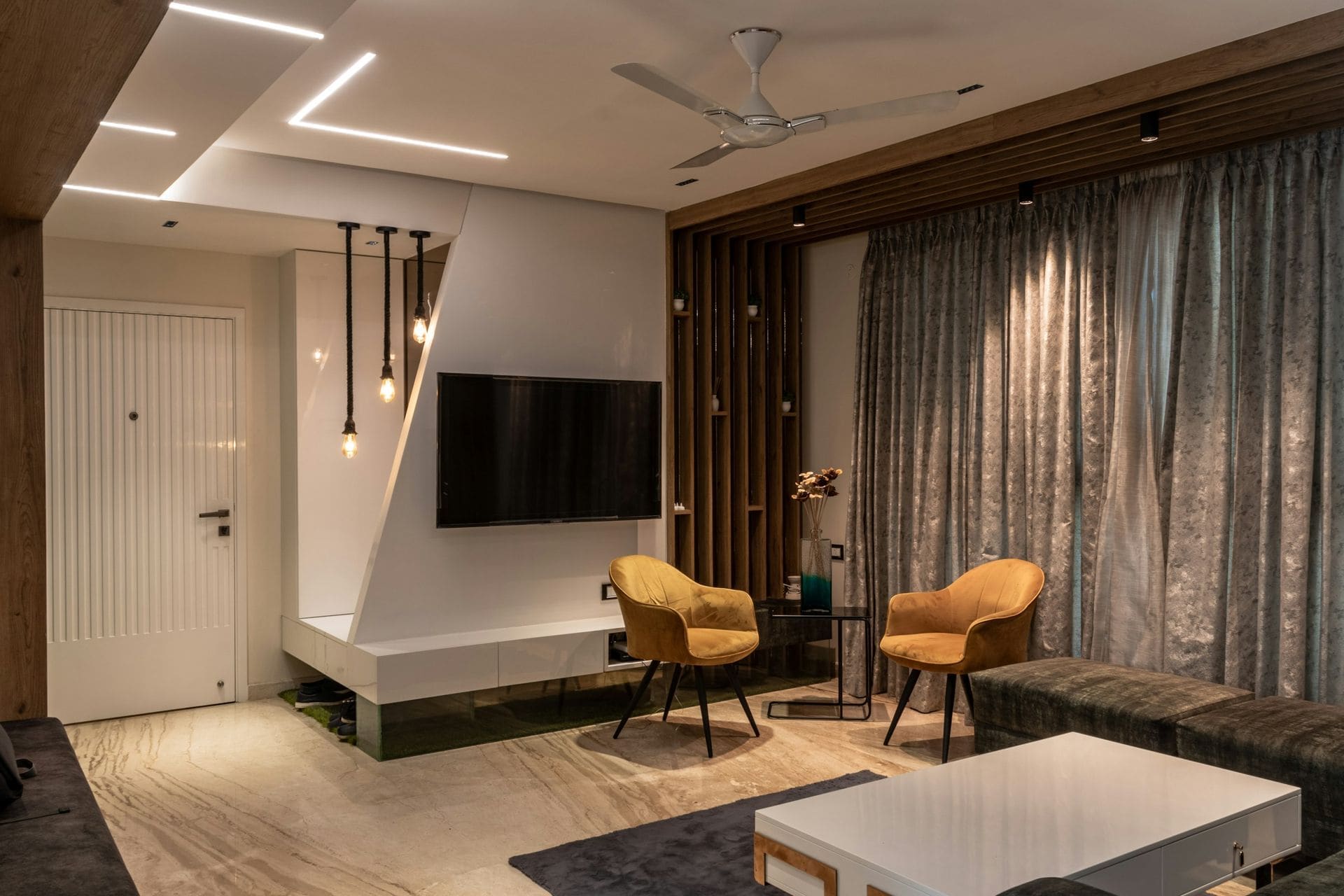 Indian living room TV wall with white floating console, wood slat partition, profile lights, ceiling fan and mustard chairs