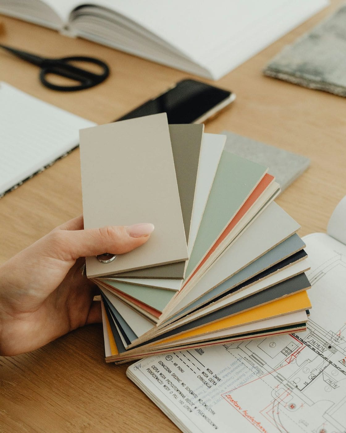 Hand holding a fan of neutral colour swatches over floor plans and an open book on a wooden desk